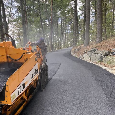 A crew member operating a yellow LeeBoy 7000 asphalt paver, laying fresh blacktop on a wooded rural driveway edged with boulders.