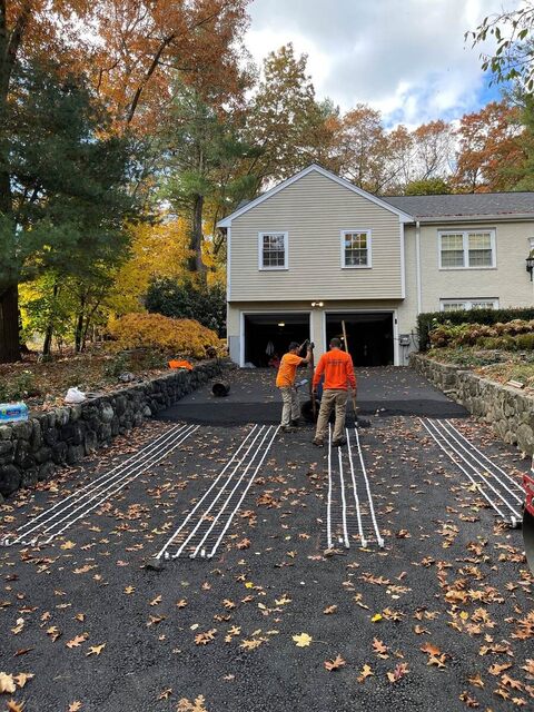 Crew laying radiant snow-melt heating cable in parallel runs across a base-coat driveway in front of a two-car garage, autumn leaves on the ground.