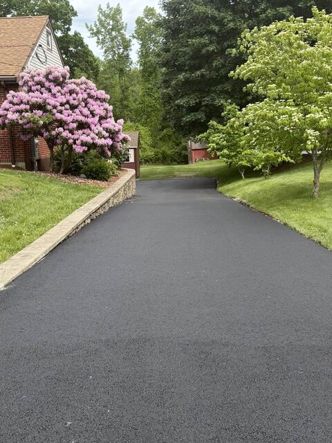 Finished black asphalt driveway curving past a brick house, edged with a low stone retaining wall and a blooming pink rhododendron.