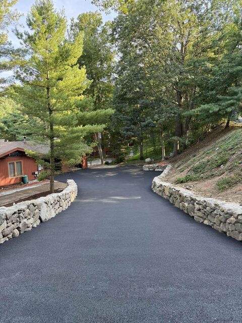 A finished curved asphalt driveway running between two new dry-stacked stone retaining walls, cut into a wooded hillside lot.