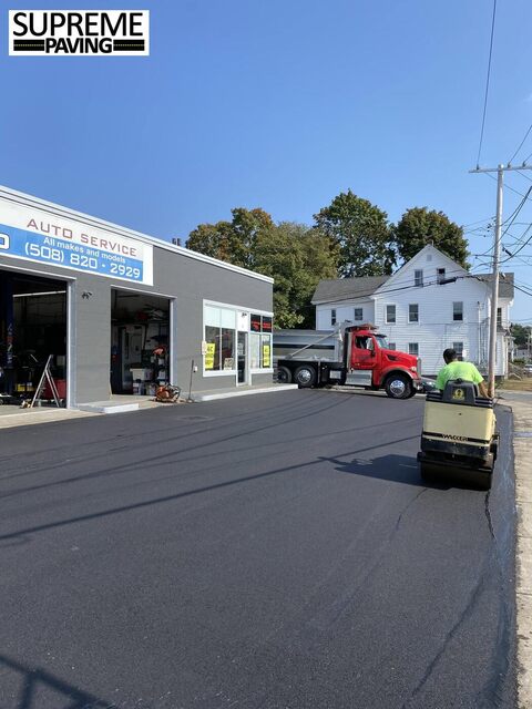 Supreme Paving's red dump truck and a crew member on a roller compactor, resurfacing the lot outside an auto-service shop.