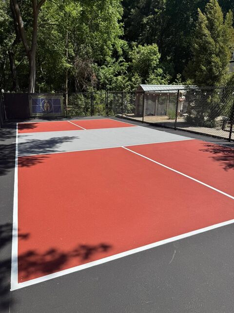 A freshly coated red-and-gray backyard pickleball court with crisp white lines, inside a chain-link enclosure.