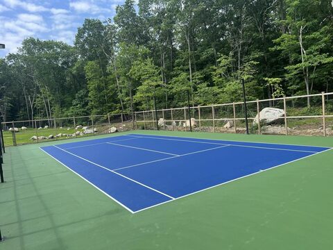 A completed blue-and-green tennis court, freshly coated and fully lined, set into a wooded clearing behind new fencing — Supreme Paving's sport-court work.